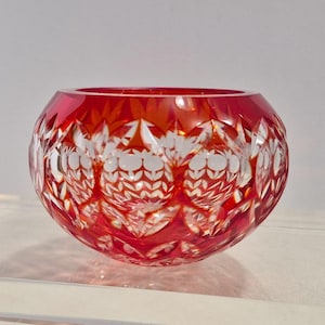 May include: A round, red glass bowl with a cut crystal design. The bowl has a wide opening and a thick base. The cut crystal pattern features a repeating floral motif. The bowl is likely used for decorative purposes.