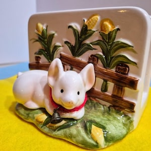 May include: A ceramic napkin holder featuring a white pig with a red collar and bell, set against a backdrop of corn stalks and a wooden fence. The base is green with corn details.