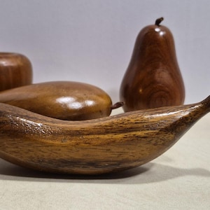 May include: A collection of wooden fruit sculptures, including a banana, pear, apple, and a mango-shaped piece. The sculptures are all crafted from a warm, brown wood, showcasing the natural grain and texture of the material.