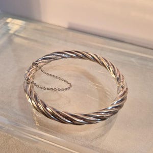 May include: A silver-colored bangle bracelet with a twisted rope design. The bracelet is round and has a clasp with a safety chain. The jewelry is displayed on a clear, reflective surface.