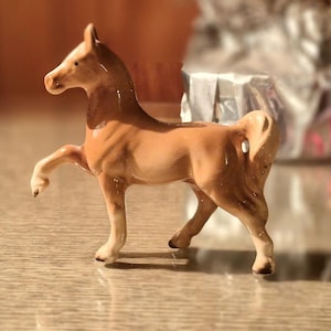 May include: A small, tan-colored horse figurine with one leg raised. The horse has a detailed mane and tail, and is posed as if in motion. The figurine is made of a glossy material and is set on a light-colored surface.