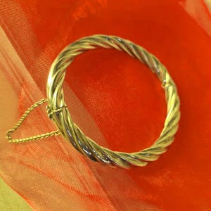 May include: A silver-toned bangle bracelet with a twisted rope design. The bracelet is circular and has a clasp closure with a safety chain. The background is a vibrant red fabric.