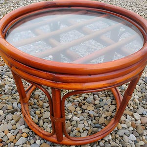 May include: A round, brown rattan side table with a clear glass top. The table has a woven pattern beneath the glass and curved legs. The table is approximately 24 inches in diameter and 20 inches tall.
