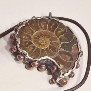 May include: A pendant featuring a polished ammonite fossil, showcasing a spiral pattern in shades of brown and tan. The fossil is set in a silver-toned metal frame with copper-colored accents, suspended from a brown leather cord.