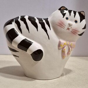May include: A ceramic cat figurine with a white base and black stripes. The cat has blue eyes, pink cheeks, and a pink bow with yellow polka dots. The figurine is a decorative item.