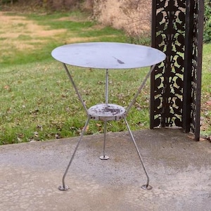 May include: A small, round metal side table with a three-legged base. The tabletop is a flat, silver disc, and the legs are thin, angled metal rods. The table has a small, circular shelf beneath the tabletop. The table is outdoors on a concrete surface.