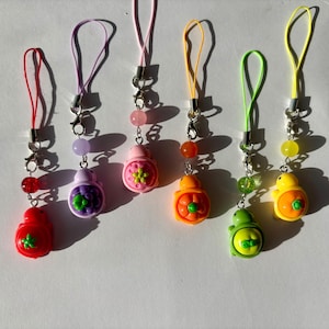 May include: A collection of six colorful phone charms. Each charm features a small, decorative container in red, purple, pink, orange, green, and yellow, with a matching cord and bead accents. The charms are designed with various floral and fruit-like details.