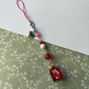 Puede incluir: Un charm con una fresa y un tarro de mermelada. El charm tiene un cordón rosa y está adornado con cuentas rosas, blancas y rojas. El tarro de mermelada es rojo con una tapa dorada y una etiqueta de fresa.