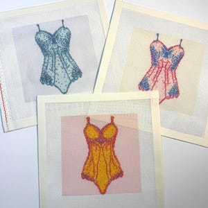 May include: Three square needlepoint canvases featuring cross-stitch designs of one-piece swimsuits. One canvas displays a light blue swimsuit, another a pink and blue swimsuit, and the third a yellow and orange swimsuit on a pink background.