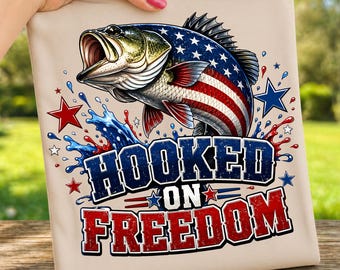 May include: A beige t-shirt featuring a detailed illustration of a largemouth bass with a patriotic design. The fish's body incorporates the American flag, and the text "HOOKED ON FREEDOM" is prominently displayed in red and blue.