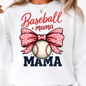 May include: White sweatshirt with a baseball-themed graphic. The design features the words "Baseball mama" in red script, a pink bow with white polka dots, a baseball, and the word "MAMA" in navy blue block letters.