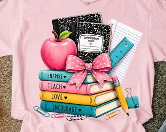 May include: A light pink t-shirt featuring a graphic of a stack of books labeled Inspire, Teach, Love, and Encourage. The design includes a pink apple, a black and white composition book, a blue ruler, a yellow pencil, and a pink polka dot bow.