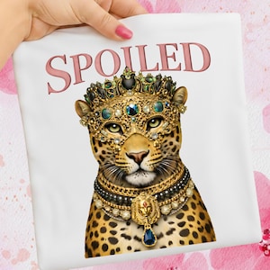 Op de afbeelding: Wit t-shirt met een illustratie van een koninklijke luipaard. De luipaard draagt een met juwelen bezette kroon, masker en ketting. Het woord "SPOILED" is in roze boven de kop van de luipaard gedrukt.
