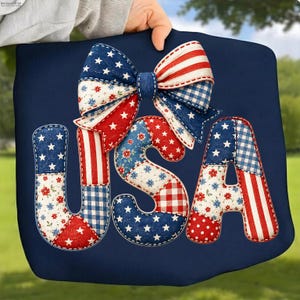Op de afbeelding: Een marineblauw kussen met het woord "USA" in patchworkletters in rood, wit en blauw. Boven de letters staat een grote strik met rode en witte strepen en blauwe sterren. Het kussen wordt door een persoon vastgehouden.