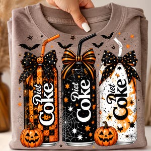 May include: A light brown t-shirt featuring a graphic of three Diet Coke cans decorated with Halloween themes, including orange and black bows, straws, spider webs, stars, bats, and jack-o'-lanterns at the base of the cans.