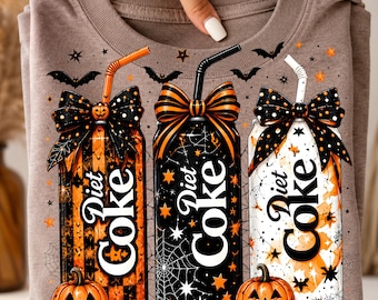 May include: A light brown t-shirt featuring a graphic of three Diet Coke cans decorated with Halloween themes, including orange and black bows, straws, spider webs, stars, bats, and jack-o'-lanterns at the base of the cans.
