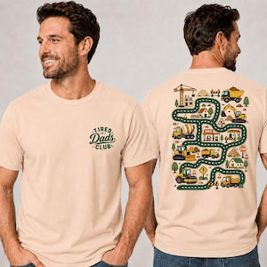 Puede incluir: Una camiseta beige con un pequeño gráfico de texto verde en la parte delantera que dice Tired Dads Club. La parte posterior de la camiseta muestra una gran ilustración colorida de una carretera sinuosa con vehículos de obra, edificios y árboles.