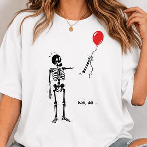 White shirt mockup featuring funny Well Shit skeleton, floating red balloon and gothic dark humor Halloween design.