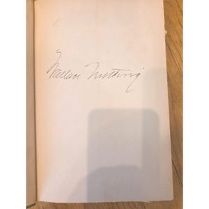 May include: An open book with a handwritten signature in black ink on a cream-coloured page. The signature appears to be a name, possibly an author's, with cursive lettering. The book is open on a wooden surface.