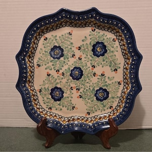 Puede incluir: Un plato cuadrado de cerámica con bordes festoneados que presenta un centro de color crema decorado con cinco flores azul oscuro y follaje verde claro. El borde muestra un patrón repetitivo de formas geométricas azules, amarillas y marrones.