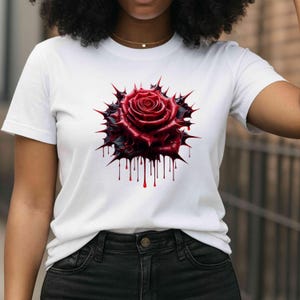 Peut inclure: T-shirt blanc avec un motif de rose rouge stylisée. La rose est entourée d'une bordure sombre et épineuse, avec des détails dégoulinants rappelant du sang. Le t-shirt est de style col rond classique.