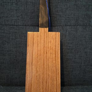 May include: A wooden cutting board with a dark handle and a blue string for hanging. The board is rectangular with a natural wood grain finish. The handle is painted black.