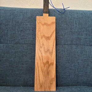 May include: A wooden paddle with a natural wood grain finish. The handle is black and has a blue string attached. The paddle is rectangular with a T-shaped handle. The wood has a light brown color.