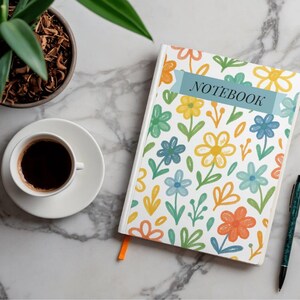 May include: A floral notebook with a blue banner that reads "NOTEBOOK" sits on a marble surface, next to a cup of coffee and a pen. The notebook has a white cover with colorful flowers. A small potted plant is in the upper left corner.