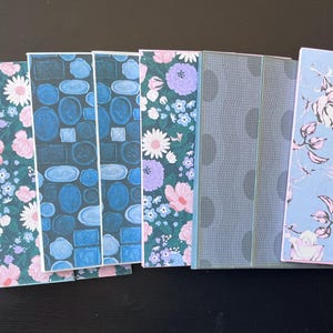 May include: A collection of eight rectangular bookmarks featuring diverse patterns including light blue floral prints, dark green backgrounds with pink and purple flowers, blue geometric shapes, a grey textured design, and a light blue rose motif.