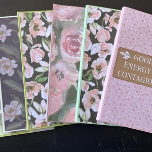 May include: A set of six rectangular greeting cards featuring various designs including butterflies, floral patterns in pink and black, and a light pink card with a brown label that reads Good Energy Is Contagious, accented with a vertical strip of burlap lace.