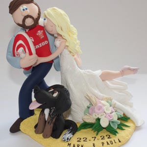 May include: A wedding cake topper featuring a bride and groom embracing, with a black and brown dog at their feet. The groom holds a rugby ball. The bride wears a white wedding dress. The base is yellow with the date "22-7-22" and names "MARK & PAULA".