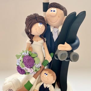 Könnte beinhalten: Hochzeitstortenaufsatz mit Braut- und Bräutigamfiguren, einem Kind und einem Wegweiser nach Limerick und Offaly. Die Braut hält einen Strauß lila und weißer Blumen. Der Bräutigam hält schwarze Skier.