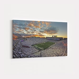 Op de afbeelding: Een panoramisch uitzicht op een vol voetbalstadion bij zonsondergang. Het veld is groen, omringd door witgeklede fans. De lucht is gevuld met oranje en grijze wolken, wat een dramatische achtergrond creëert.