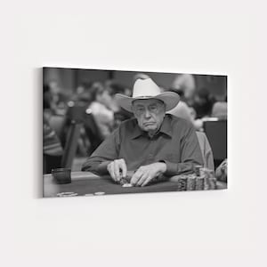 May include: Black and white photograph of an older person wearing a cowboy hat, seated at a poker table. The person is holding playing cards and surrounded by poker chips. The background is blurred, suggesting a casino setting.