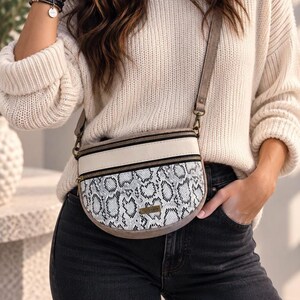 May include: A crescent-shaped crossbody bag featuring a white and black snakeskin pattern on the lower section, a solid cream-coloured upper panel, and two black zippered compartments with antique brass-toned hardware and an adjustable shoulder strap.