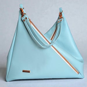 May include: A pale blue handbag with a distinctive triangular design. It has a diagonal zip with a white interior and rose gold-coloured hardware. The handles are pale blue with rose gold accents. A small, rectangular gold-coloured label is on the front.