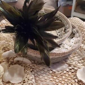 May include: Two woven baskets with lace trim, one nested inside the other, are adorned with a cluster of dark green feathers. The baskets sit on a cream-colored, open-weave fabric, with white petals scattered around.
