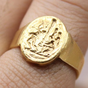 May include: A gold ring with an oval face, displaying a detailed relief design. The ring is worn on a finger, highlighting its polished finish and intricate artwork. The ring is from Classi finejewelry.