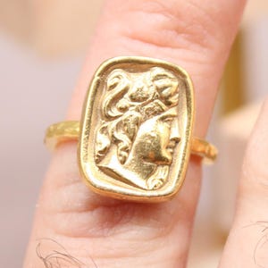 May include: A gold-colored ring featuring a rectangular face with a detailed relief of a classical figure. The ring is worn on a finger, showcasing the intricate design and craftsmanship of the jewelry.