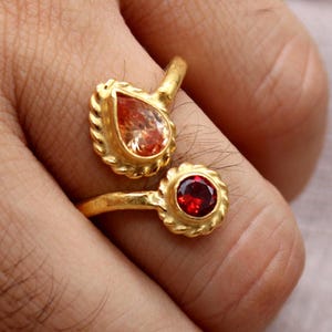 Peut inclure: Bague en or avec deux pierres précieuses, une en forme de goutte d'eau orange clair et l'autre ronde rouge. La bague est portée au doigt. Le logo Classi finejewelry est visible en bas à gauche. Bijou.