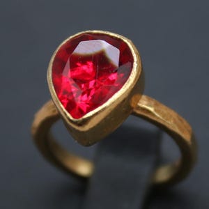 Op de afbeelding: Een gouden ring met een peervormige, levendige rode edelsteen. De ring heeft een getextureerde gouden band en wordt gepresenteerd op een zwarte standaard. De afbeelding bevat de tekst "Classi finejewelry" in een gouden logo.
