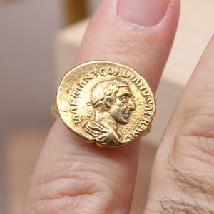 May include: A gold coin ring featuring a profile of a person with text around the edge. The ring is on a finger, with a blurred background. The ring is likely made of gold.