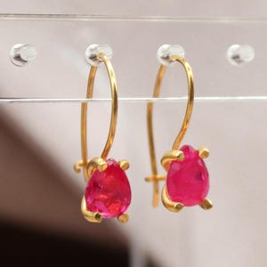 Könnte beinhalten: Ein Paar goldfarbene Ohrhänger mit leuchtend roten, tropfenförmigen Edelsteinen. Die Ohrringe werden auf einem transparenten Schmuckständer präsentiert. Das Logo "Classi finejewelry" ist unten links sichtbar.