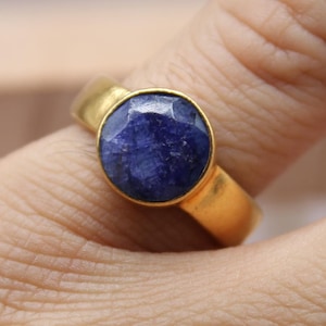 May include: A gold ring with a large, round, deep blue gemstone. The ring is set in a simple, classic style, with a smooth, polished band. The gemstone is faceted, catching the light and creating a sparkling effect.