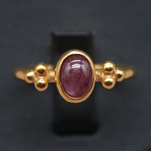 May include: A gold ring featuring a maroon oval gemstone. The ring has a delicate design with small, round accents on either side of the gemstone. The ring is displayed on a black surface. The logo "Classi finejewelry" is in the lower left corner.