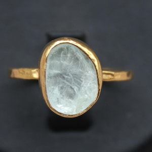 May include: A gold ring featuring a light blue, irregularly shaped gemstone. The gemstone is set in a gold band, creating a simple yet elegant design. The ring is displayed against a dark background.