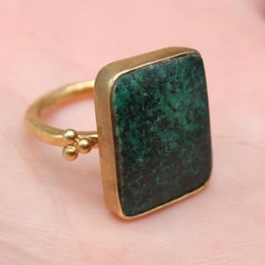 May include: A gold-toned ring featuring a rectangular, dark green stone. The ring has a simple band with a small cluster of three beads on each side of the stone. The ring is displayed on a person's palm.