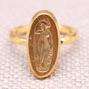 May include: A gold ring with an oval face, featuring a carved figure. The ring has a hammered texture and is set against a light background. The ring is a piece of fine jewellery.