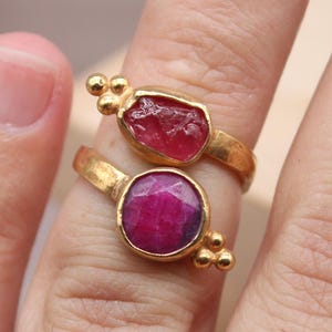 May include: Two gold rings with ruby gemstones. One ring features a raw-cut ruby, while the other has a round, polished ruby. The rings are on a finger, showcasing the jewelry's design and color. The rings are from Classi finejewelry.