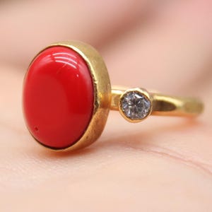 Può includere: Anello d'oro con una grande gemma rossa brillante di forma ovale. Una piccola gemma rotonda e trasparente è incastonata sulla fascia. L'anello è tenuto in una mano, con il logo "Classi finejewelry".